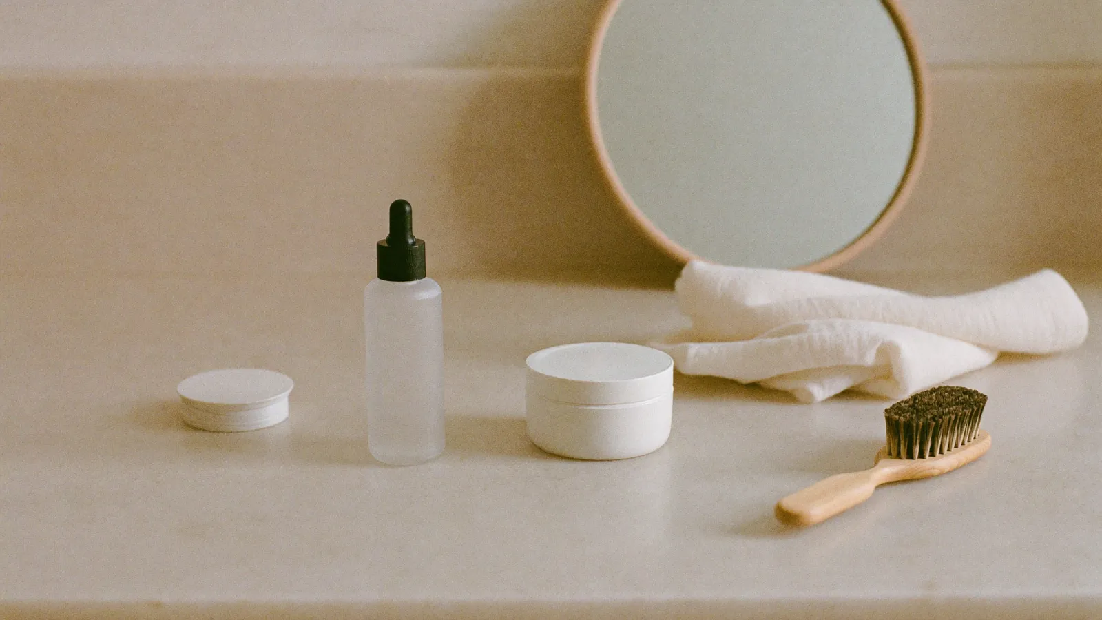 Minimalistische Morgenszene mit unbeschrifteter Glasflasche, Cremetiegel, Naturborstenbürste und Leinentuch auf heller Steinfläche.