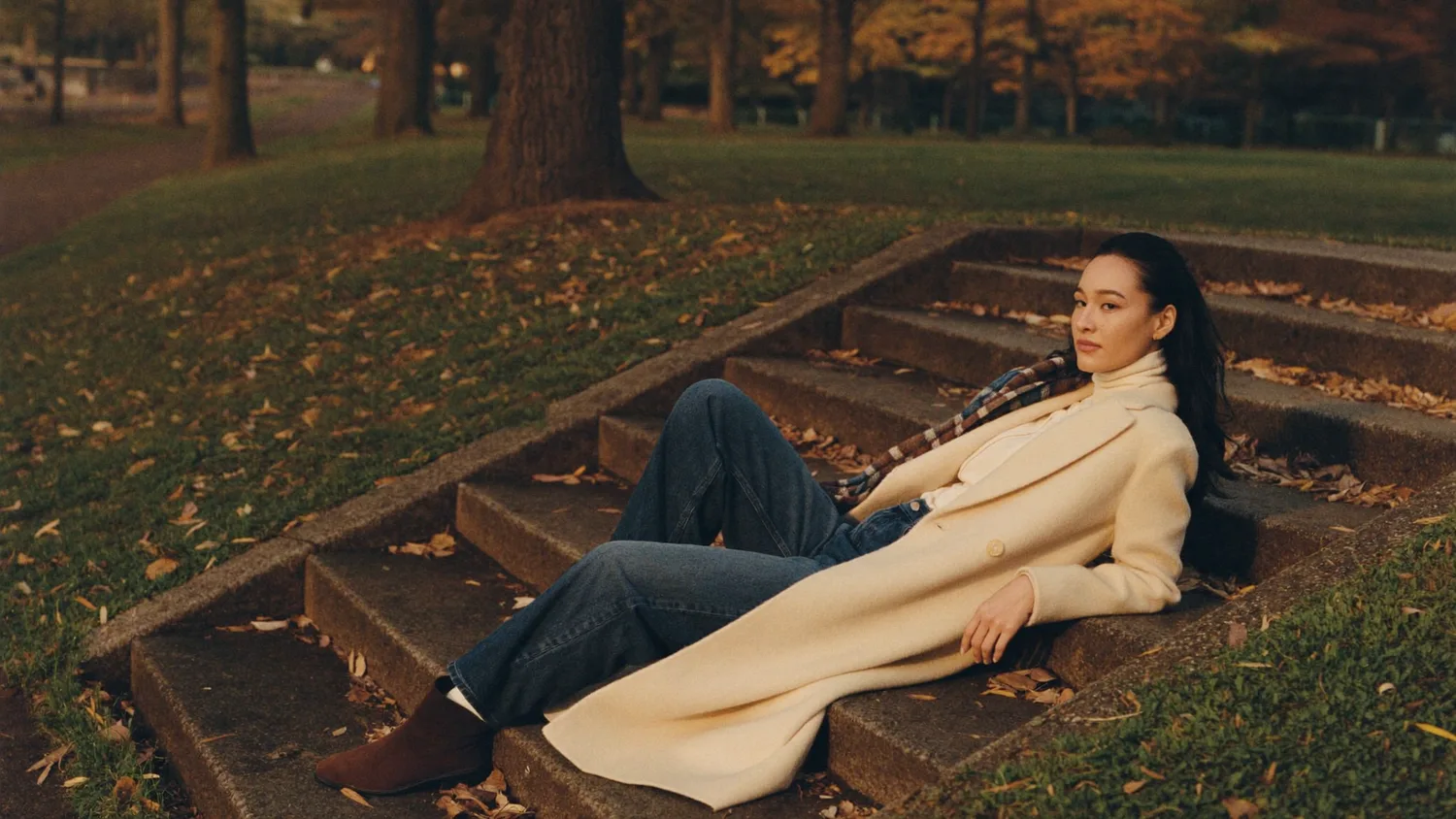 Frau auf Steinstufen in einem Park im Oktoberlicht, in Rollkragenpullover, offenem Wollmantel und dunkler Jeans.